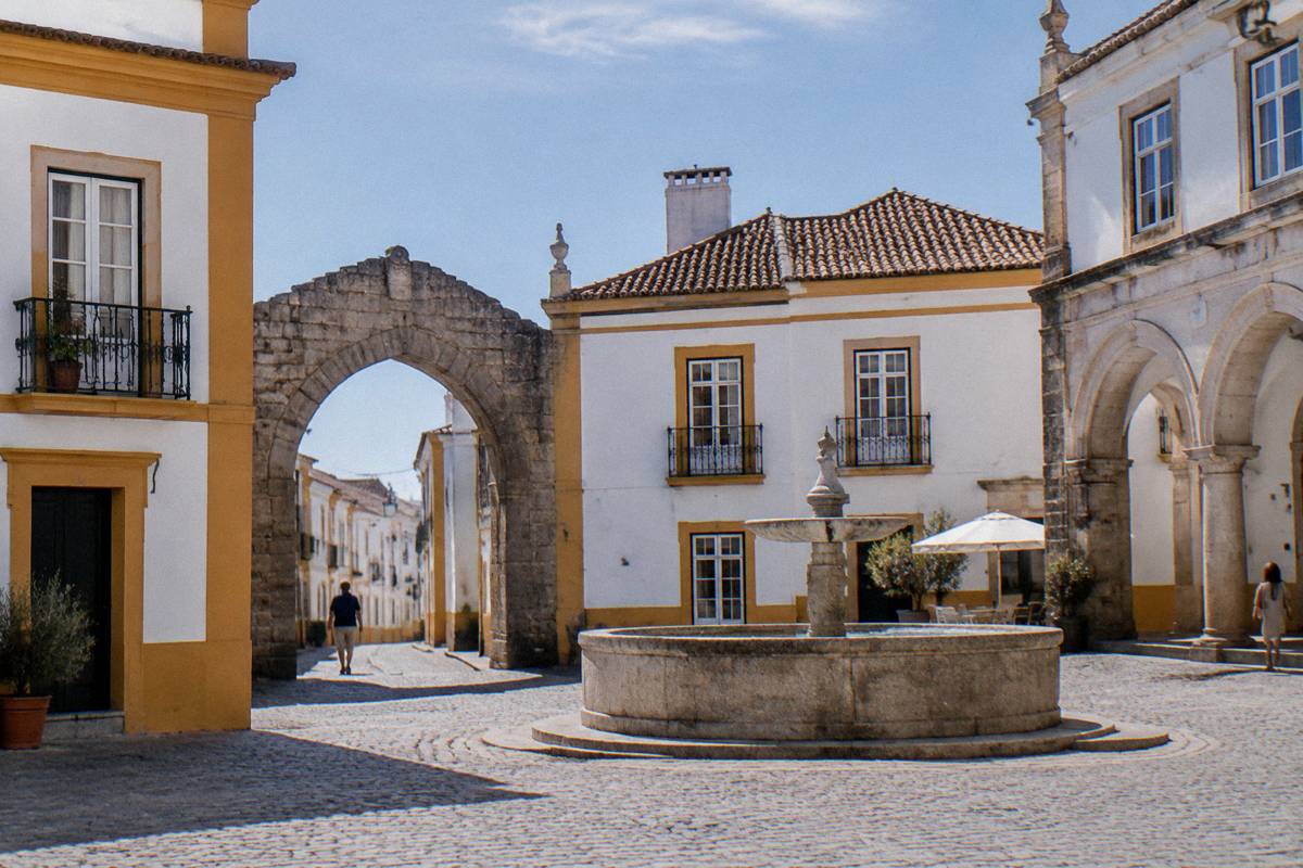 A small irregular square of whitewashed houses trimmed in ochre around a plain round stone fountain