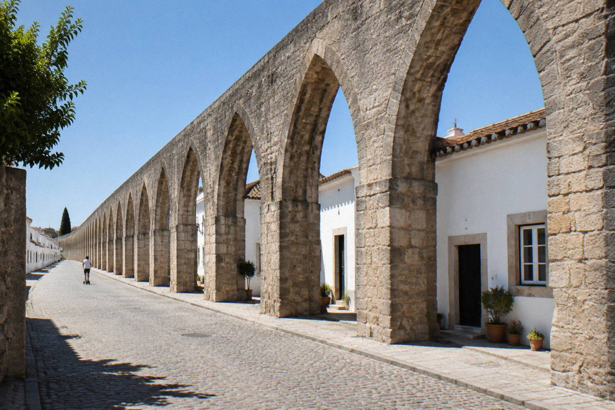 A very long stone aqueduct of pointed arches with small houses tucked beneath it and a child on a scooter far off