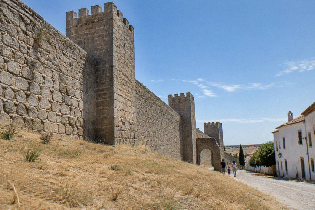 A stretch of massive granite town wall with square towers above dry grass and a family walking far from the camera