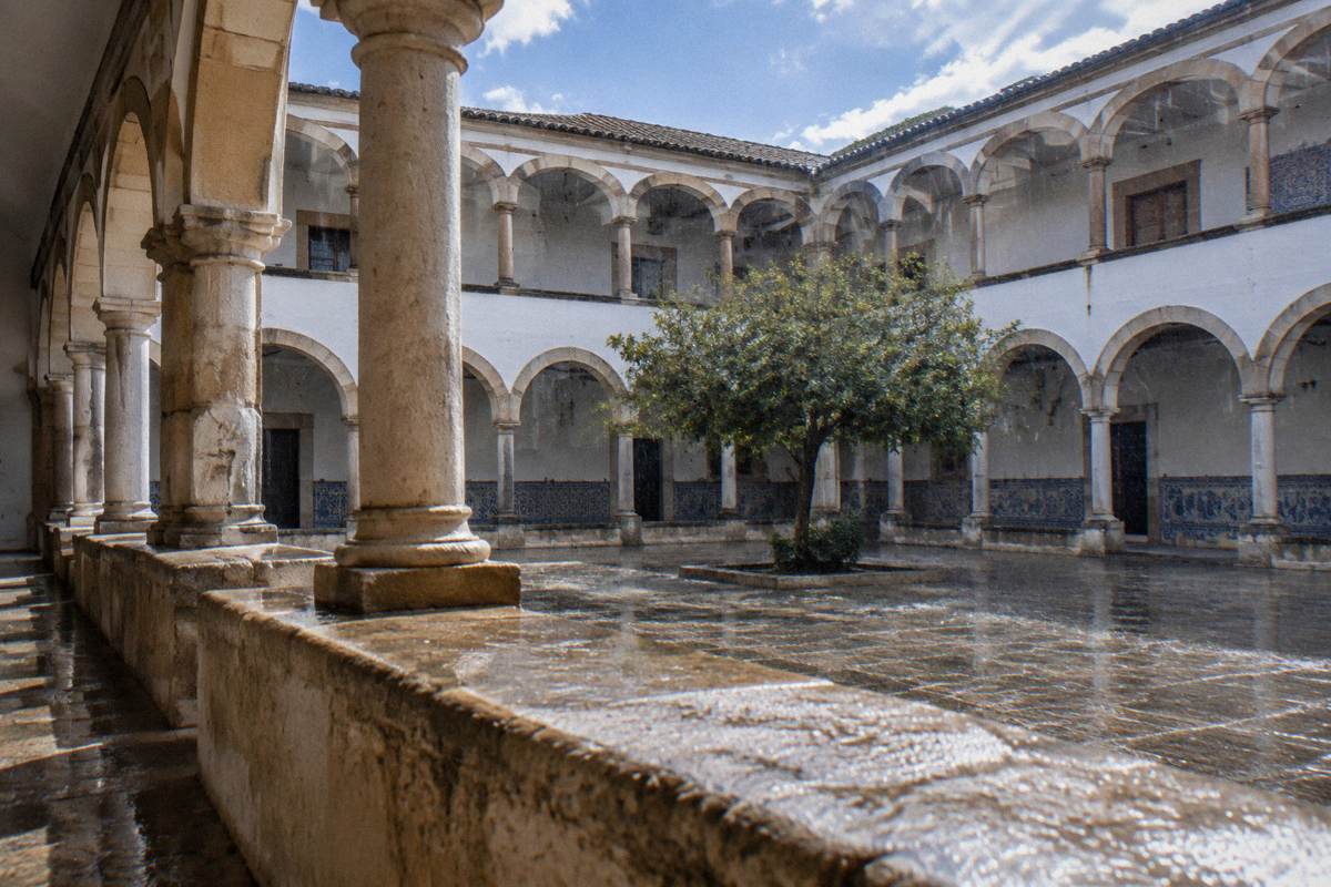 A covered cloister gallery of whitewashed walls and stone arches with rain falling on the paved court