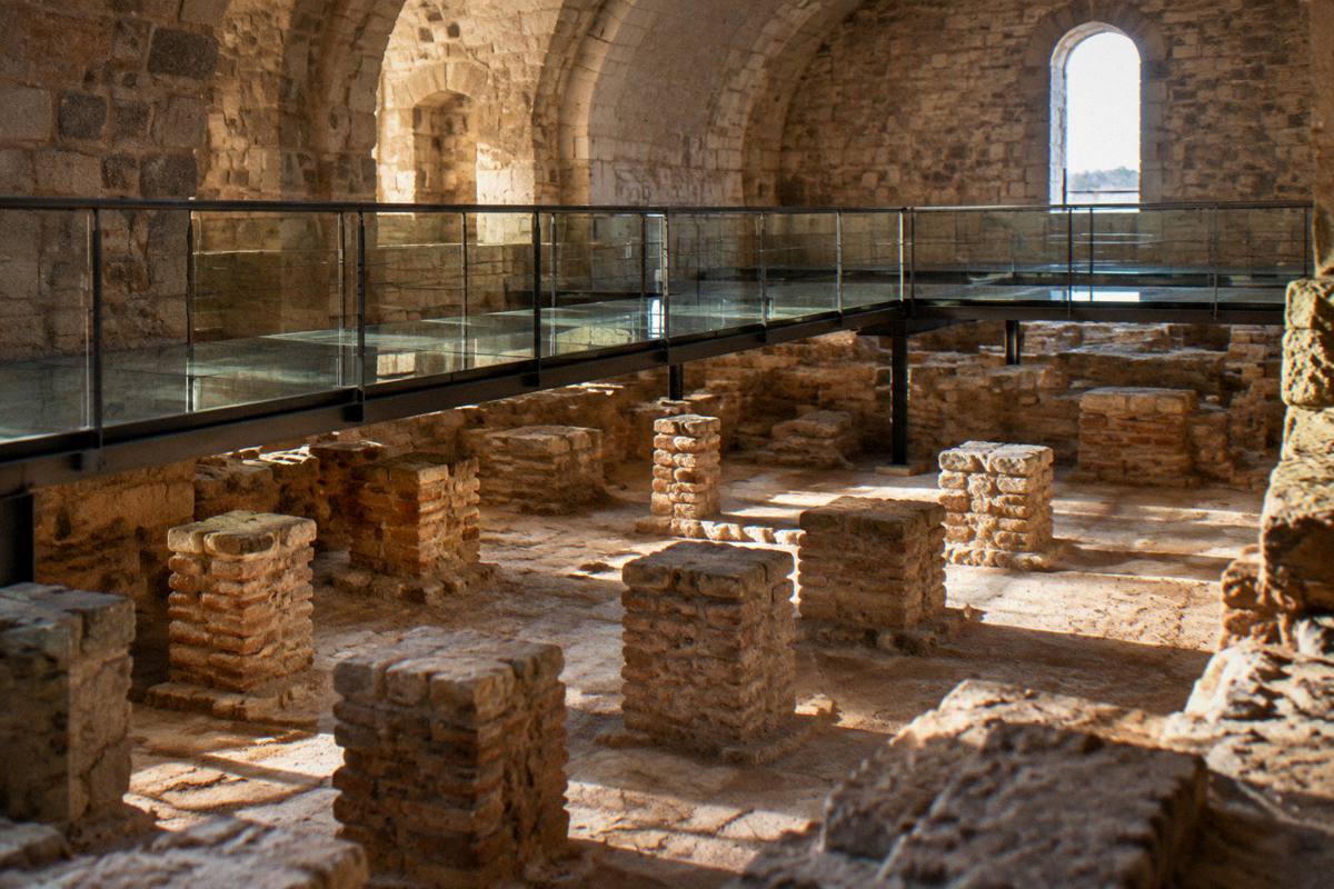 A lit excavated floor of low stone and brick pillars beneath a modern walkway in a vaulted stone room
