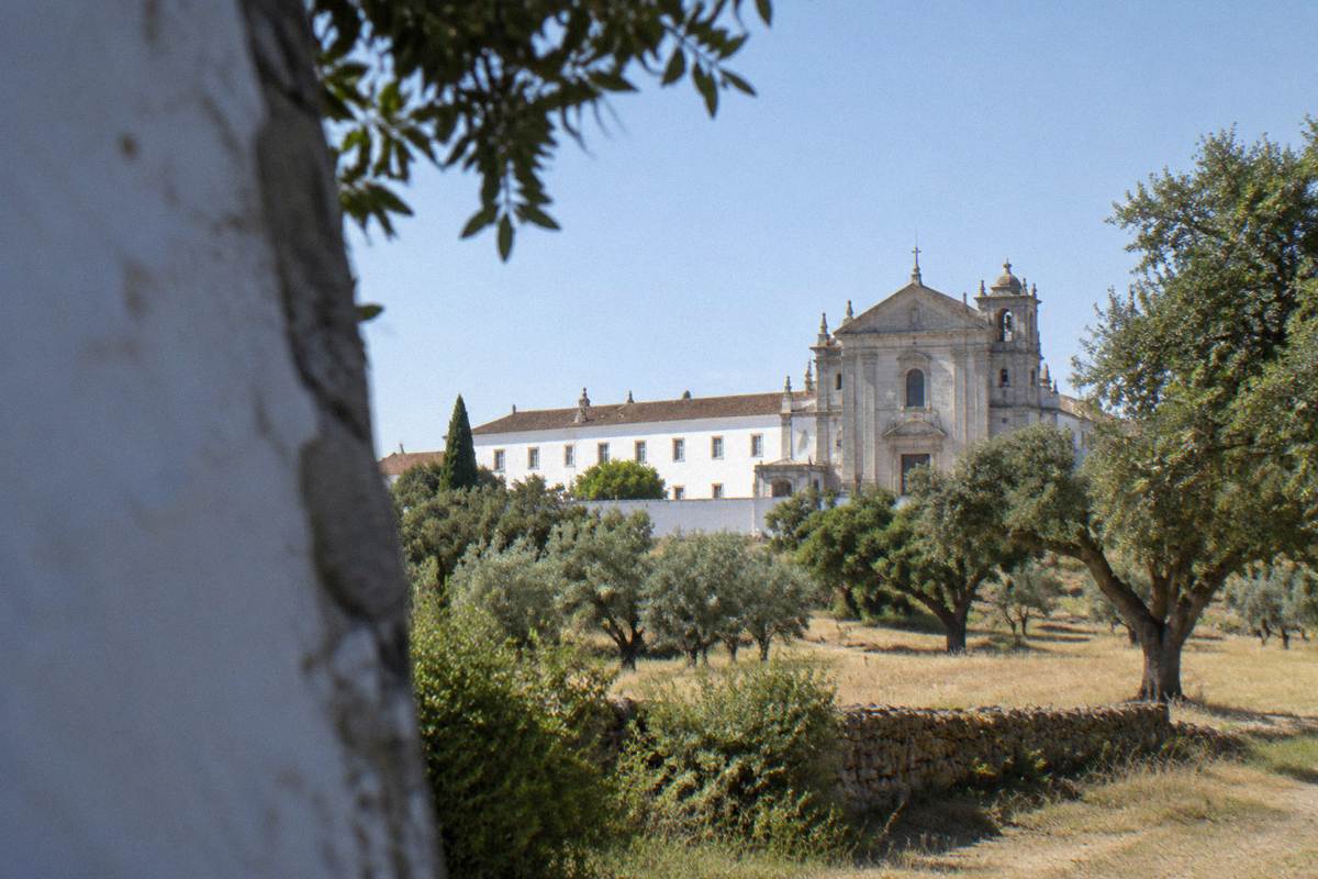 A plain whitewashed monastery with a stone church front among olive trees and cork oaks on open farmland
