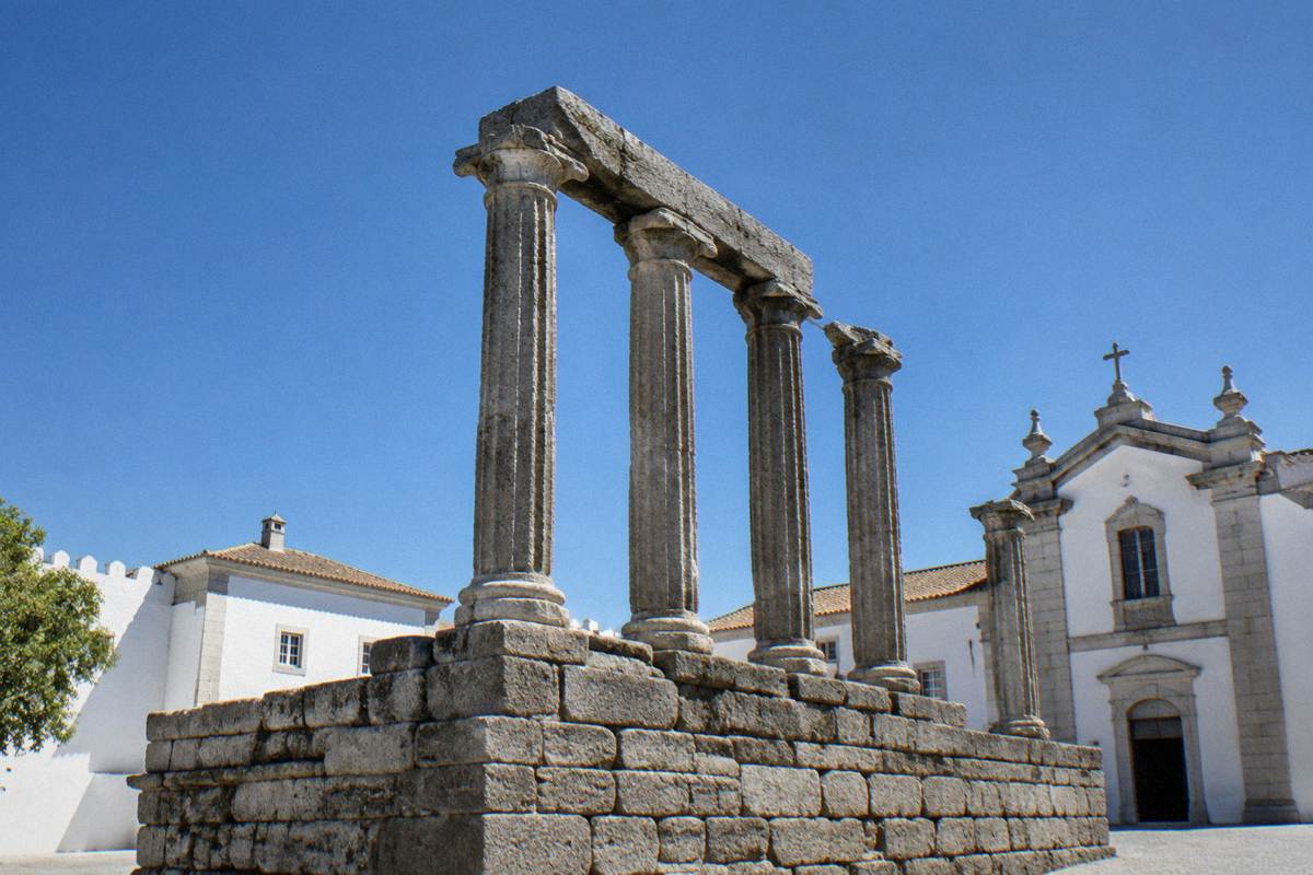 A ruined Roman temple of granite columns on a stone platform seen on a clear day with whitewashed buildings behind