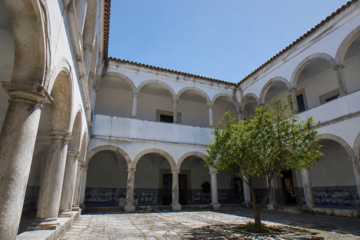 A calm cloister of whitewashed walls and pale stone arches around a paved court with blue and white tiled panels in shade