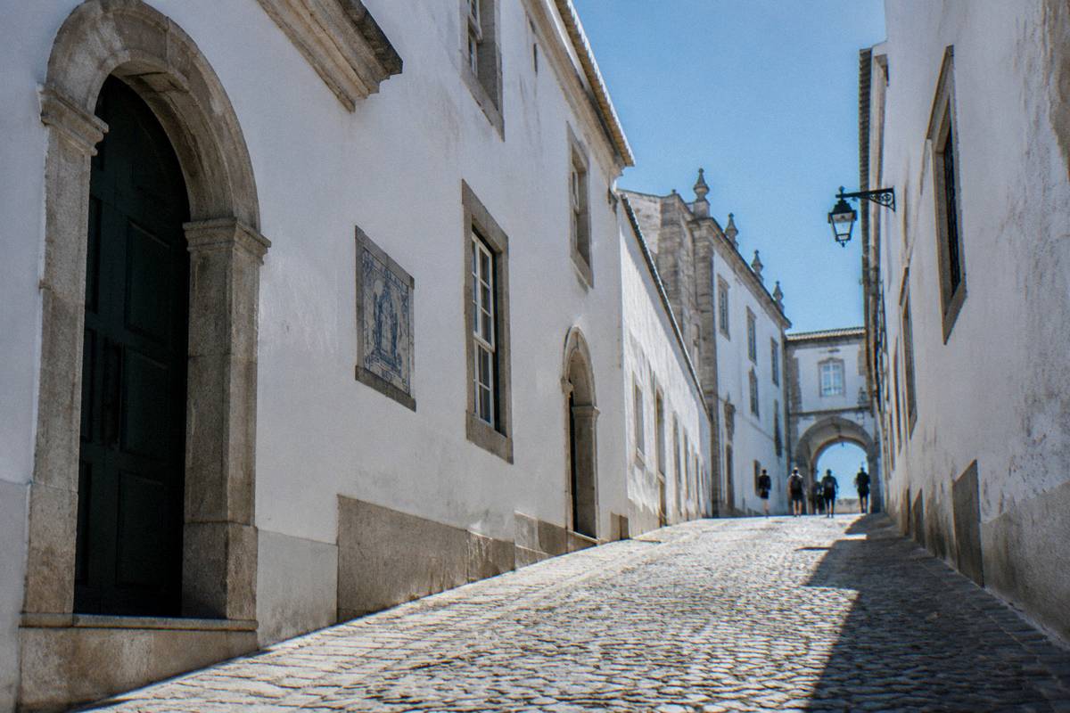 A whitewashed street of pale stone college buildings with arched doorways and students far off