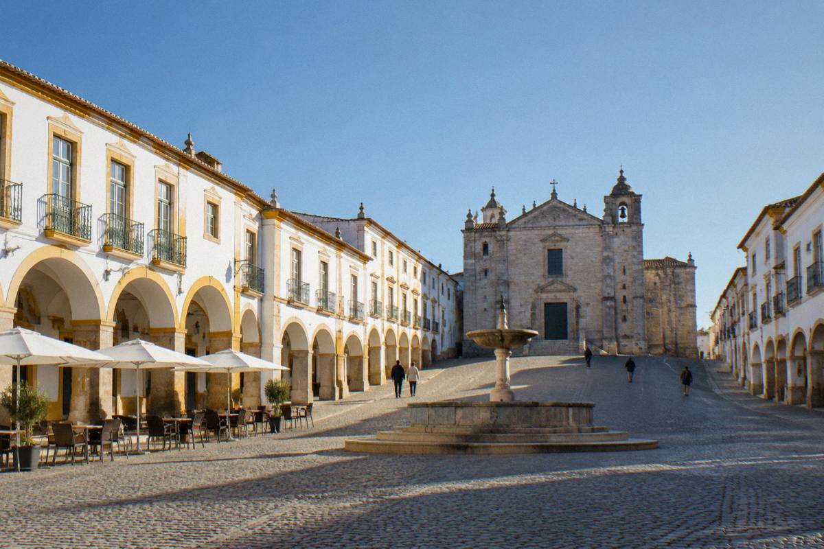 Early morning in a broad sloping square of whitewashed arcades with a plain stone fountain and a few distant early walkers
