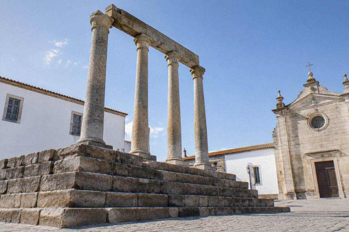 Afternoon on a quiet paved terrace where slender granite columns stand on a stepped stone platform beside whitewashed walls