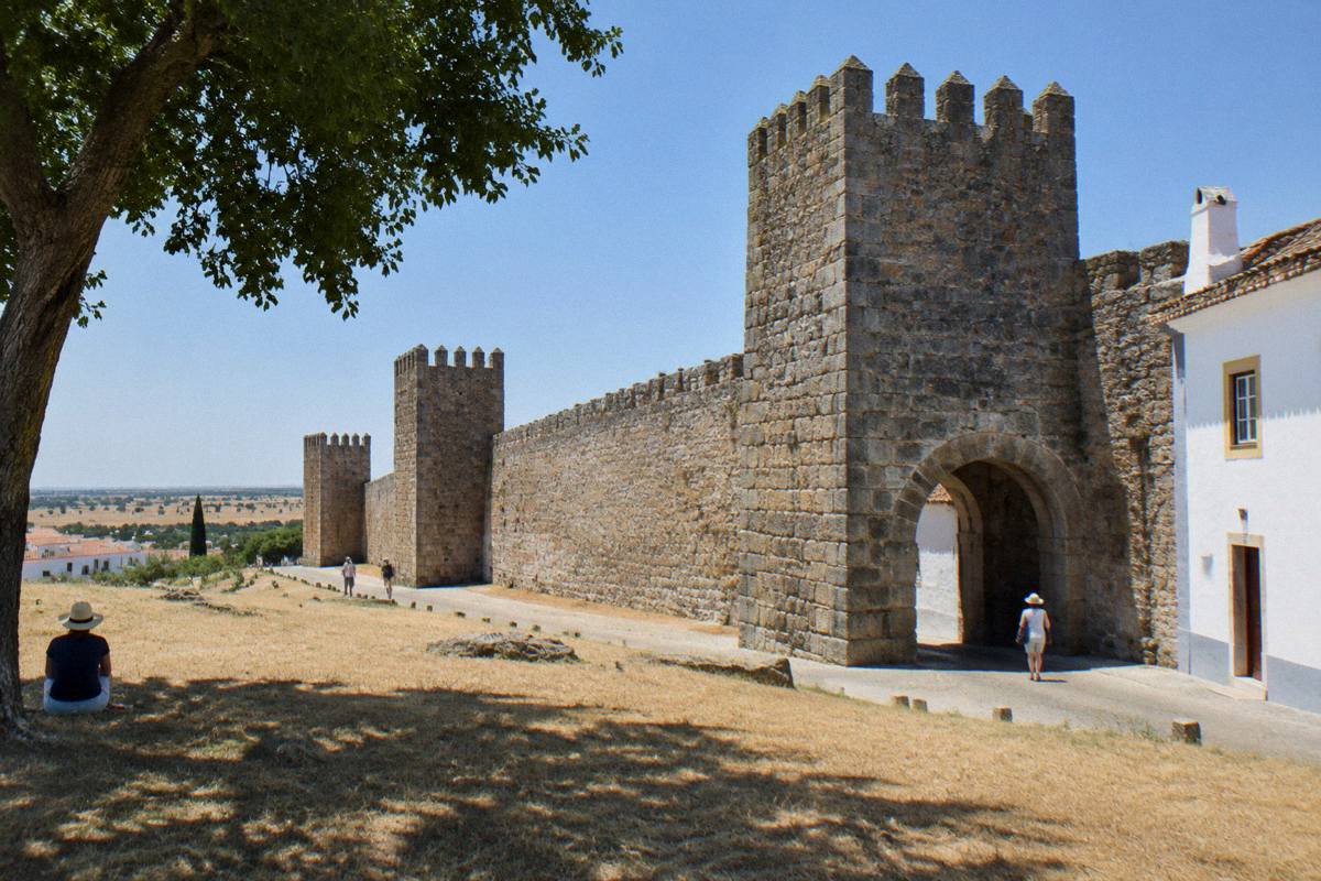 A stretch of granite town wall in hard white midday sun with dry golden grass and the shade of a tree