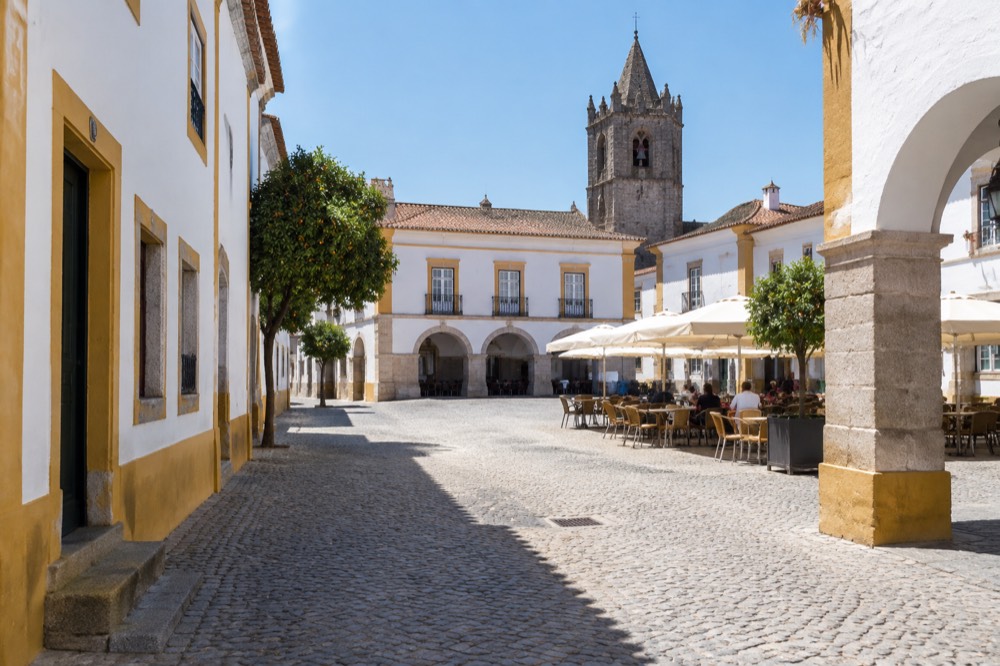 A sunlit square in Évora with white buildings, arcades and café tables set out on the cobbles