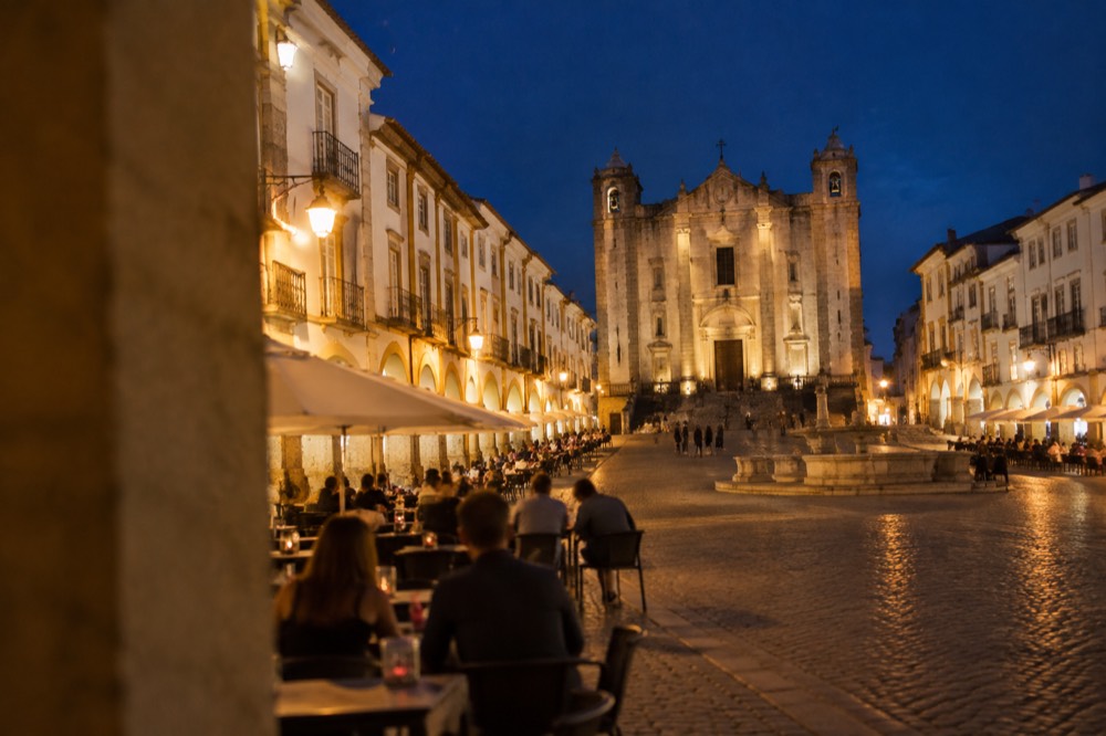 Évora's Praça do Giraldo lit up in the evening with people at outdoor tables.
